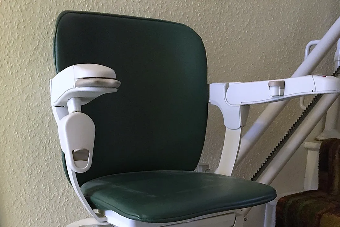 Siège vert d'un monte-escalier avec ses accoudoirs blancs, devant le rail à crémaillère qui longe l'escalier